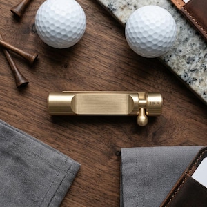 May include: A gold-colored golf ball marker with the initials "J.R.S." engraved on the side. Two white golf balls, wooden golf tees, and a leather wallet are also visible on a wooden surface.