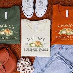 May include: Three t-shirts in green, cream, and orange, each with a pumpkin farm design. The design includes the text "Hagrid's Pumpkin Farm" and the year "1958". White trainers, a brown purse, and blue jeans are also visible.
