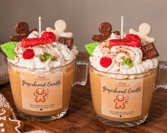 May include: Two glass mugs filled with tan wax, topped with white whipped wax, a brown gingerbread man, a white gingerbread man, a red candy cane, a green leaf, a red flower, and a chocolate square. Labels read Lovely Candles Gingerbread Candle.