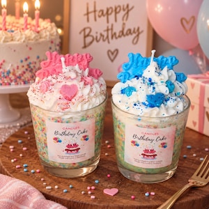 Puede incluir: Dos velas de cristal con forma de tarta de cumpleaños con cobertura de cera batida blanca, una con un adorno rosa de 'Happy Birthday' y un corazón, y la otra con un adorno azul. Ambas tienen etiquetas que dicen 'Birthday Cake' y chispas de colores.