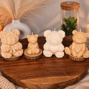 Puede incluir: Cuatro velas con forma de oso dispuestas sobre una tabla de madera. Dos velas tienen una superficie texturizada similar a rosas, una con un lazo. Dos velas tienen un acabado liso, una sosteniendo un corazón y otra sentada. Cada vela tiene una mecha blanca.