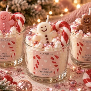 Puede incluir: Tres velas de cristal cubiertas con cera batida, una figura de hombre de jengibre y un bastón de caramelo. Cada etiqueta rosa lleva el texto CANDLE COZY CHRISTMAS MOOD con dibujos de bastones de caramelo y pequeñas esferas de color rosa.