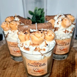 May include: Three cinnamon roll candles in glass jars. Each candle is decorated with white frosting, caramel, and a chocolate bar. The jars have labels that read "Lovely Candles Cinnamon Rolls".