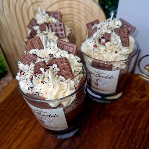 Puede incluir: Dos velas de cristal rellenas de cera marr&oacute;n oscuro, cubiertas con cera blanca batida que parece nata, decoradas con trocitos de chocolate, migas de galleta y virutas de colores. Cada etiqueta lleva el texto Hot Chocolate.