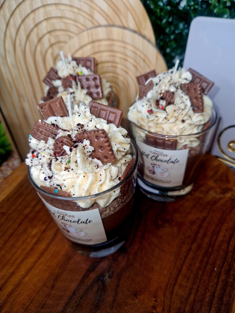 Puede incluir: Dos velas de cristal rellenas de cera marr&oacute;n oscuro, cubiertas con cera blanca batida que parece nata, decoradas con trocitos de chocolate, migas de galleta y virutas de colores. Cada etiqueta lleva el texto Hot Chocolate.