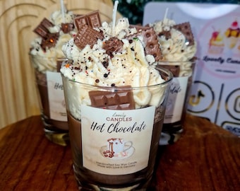 May include: Three glass candles resembling hot chocolate, topped with whipped cream, chocolate pieces, and sprinkles. Each candle has a label that reads "Hot Chocolate" and "Lovely Candles". The candles are on a wooden surface.