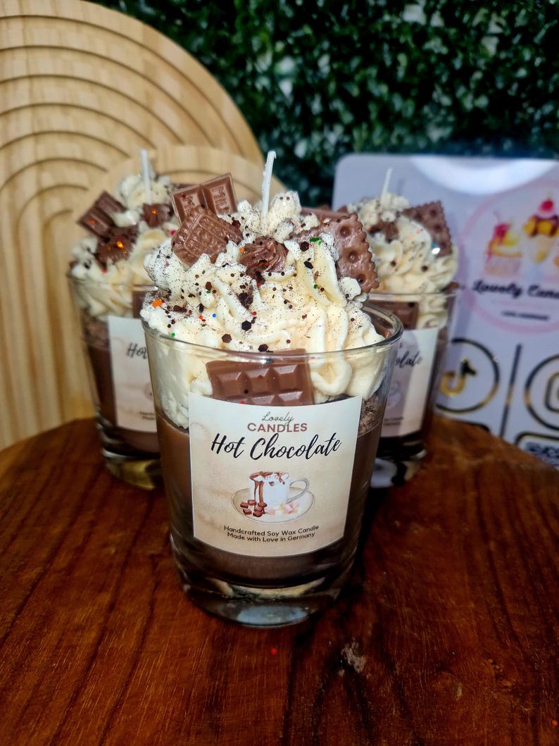 Puede incluir: Una vela en vaso con base marr&oacute;n y cobertura de cera batida blanca, decorada con formas en miniatura de barritas de chocolate y galletas. La etiqueta dice "Lovely CANDLES Hot Chocolate" con una ilustraci&oacute;n de una taza y el texto "Handcrafted Soy Wax Candle Made with Love in Germany".