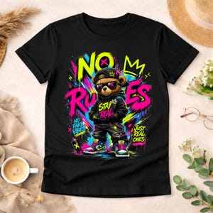 May include: Black t-shirt featuring a cartoon bear wearing sunglasses, a cap, and a hoodie. The bear is surrounded by graffiti-style text that reads "NO RULES", "STAY REAL", and "JUST REAL ONES". The design includes vibrant colors like pink, yellow, and blue.