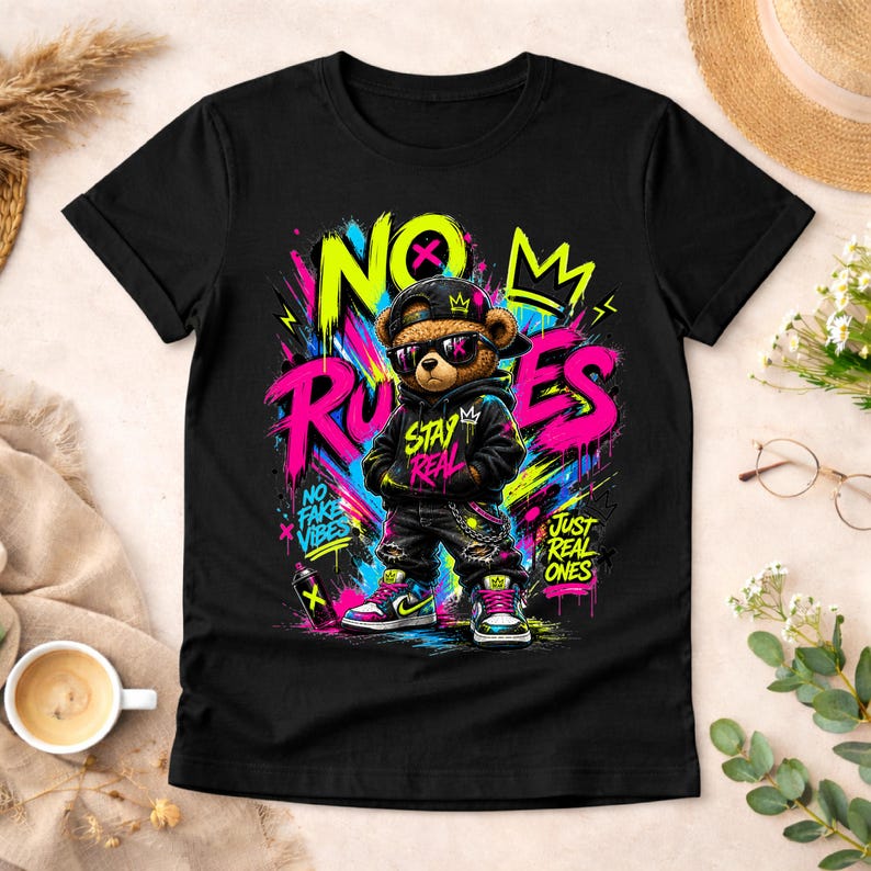 May include: Black t-shirt featuring a cartoon bear wearing sunglasses, a cap, and a hoodie. The bear is surrounded by graffiti-style text that reads "NO RULES", "STAY REAL", and "JUST REAL ONES". The design includes vibrant colors like pink, yellow, and blue.