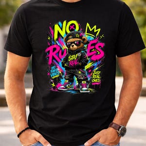 May include: Black t-shirt featuring a cartoon teddy bear wearing sunglasses, a black hoodie, and sneakers. The bear is in front of graffiti-style text that reads "NO RULES" and "STAY REAL". The shirt has a vibrant, colorful design.