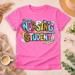 May include: A vibrant pink t-shirt with the text "NURSING STUDENT" in a colourful design. The design includes medical symbols such as a stethoscope, syringe, and thermometer, ideal for nursing students.