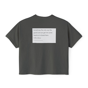 May include: A dark gray t-shirt with a white rectangle on the back. The rectangle contains a quote: "Sometimes the only way the good Lord can get into some hearts is to break them." The quote is attributed to Fulton J. Sheen.