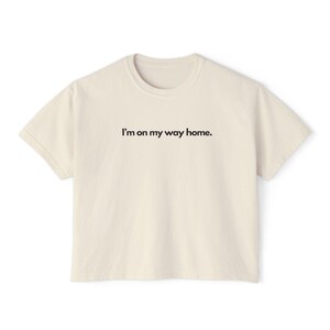 May include: A cream-coloured t-shirt with short sleeves and a crew neck. The shirt has the text "I'm on my way home." printed in black on the front.