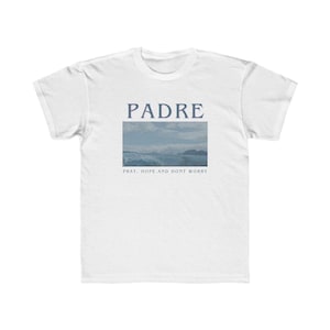 May include: White t-shirt with a blue graphic of a seascape and the text "PADRE" above it. The text below the image reads "Pray, Hope and Don't Worry".