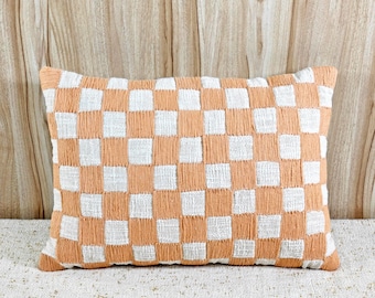 May include: Rectangular decorative pillow with a checkerboard pattern in peach and white. The pillow is made of a textured fabric and is placed on a neutral-colored surface. The background is a light wood panel.