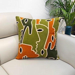 May include: Square cushion with an abstract face design. The cushion features a textured, embroidered pattern in shades of green, orange, and black, with white outlines. The cushion is placed on a white sofa.