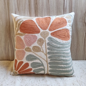 Puede incluir: Cojín decorativo cuadrado blanco con diseño floral bordado. El diseño presenta flores y hojas de color naranja, rosa y verde azulado. El cojín está hecho de una tela suave y texturizada, perfecto para añadir un toque de decoración inspirada en la naturaleza.