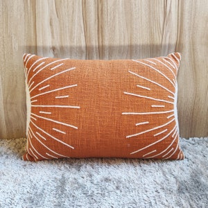 May include: A rectangular orange cushion with a white sunburst design. The cushion is made of a textured fabric and has a subtle sheen.