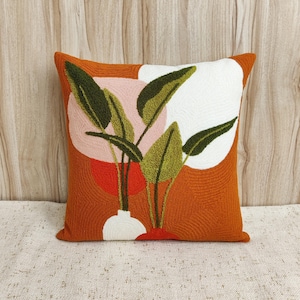 May include: Orange and white throw pillow with an embroidered design of green leaves and a vase. The pillow has a textured fabric.