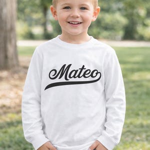May include: A child wearing a white long-sleeved top with the name "Mateo" in black script. The child is smiling and wearing blue jeans. The background is a blurred outdoor scene.