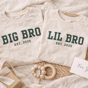 Puede incluir: Dos camisetas de cuello redondo color blanco roto con letras de estilo universitario en verde oscuro. Una camiseta muestra el texto BIG BRO EST. 2025 y la otra LIL BRO EST. 2025. Las camisetas est&aacute;n dispuestas con un mordedor de madera y una caja de regalo.