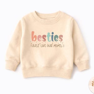 以下が含まれることがあります： パステルカラーの「besties」の文字が入ったクリーム色の子供用スウェットシャツ。「besties」の下には、「just like our moms」という手書き風のフォントで書かれたフレーズがあります。小さな円形のロゴが右下にあります。