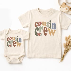 May include: A cream-coloured baby onesie and t-shirt set. Both items display the text "Cousin Crew" in colourful, retro-style lettering with a heart. A small wooden toy camera and dried flowers are also in the image.