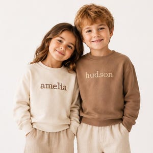 Op de afbeelding: Twee sweatshirts met lange mouwen en ronde hals, één in crème en één in bruin, beide met geborduurde tekst op de borst. Het crèmekleurige sweatshirt toont de naam amelia, terwijl het bruine sweatshirt de naam hudson toont. Beide worden gedragen met wijde beige broeken en witte sneakers.