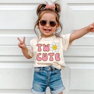 May include: A young child wearing a cream-colored t-shirt with the words "TWO CUTE" in pink and a daisy graphic. The child is also wearing blue denim shorts, sunglasses, and tan shoes. The child is smiling and making a peace sign with their fingers.