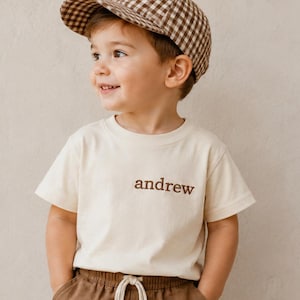 Puede incluir: Una camiseta de manga corta color crema con el nombre andrew bordado en hilo marrón en el pecho, combinada con una gorra de béisbol de cuadros vichy marrones y blancos y pantalones marrones con cordón blanco en la cintura.