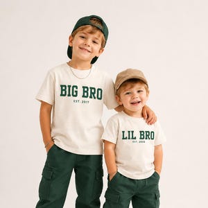 Puede incluir: Dos camisetas de manga corta color blanco roto con letras verde oscuro. Una dice BIG BRO EST. 2017 y la otra LIL BRO EST. 2025. Se combinan con pantalones cargo verde oscuro, zapatillas blancas y gorras de b&eacute;isbol en verde oscuro y color canela.