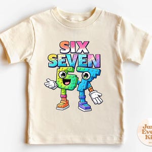 Può includere: T-shirt color crema con la scritta "SIX SEVEN" in un gradiente arcobaleno. Sotto, personaggi dei cartoni animati dei numeri 6 e 7, ognuno con braccia, gambe e facce sorridenti. La maglietta è per bambini, ideale per un compleanno.