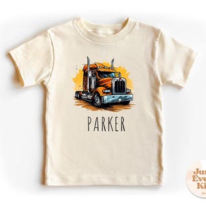 Puede incluir: Camiseta de manga corta color crema con una vibrante ilustración de un camión naranja y marrón. El nombre "PARKER" está impreso en negro debajo. La camiseta está diseñada para niños.