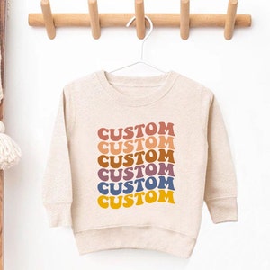 K&ouml;nnte beinhalten: Ein beiger Langarmsweatshirt mit dem Wort "CUSTOM", das sechsmal in einem Regenbogen aus Farben wiederholt wird, darunter Rot, Orange, Gelb, Gr&uuml;n, Blau und Lila.