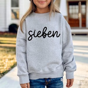 May include: A light gray sweatshirt with the word "sieben" in black script. The sweatshirt has long sleeves and a crew neck. The person is wearing blue jeans.