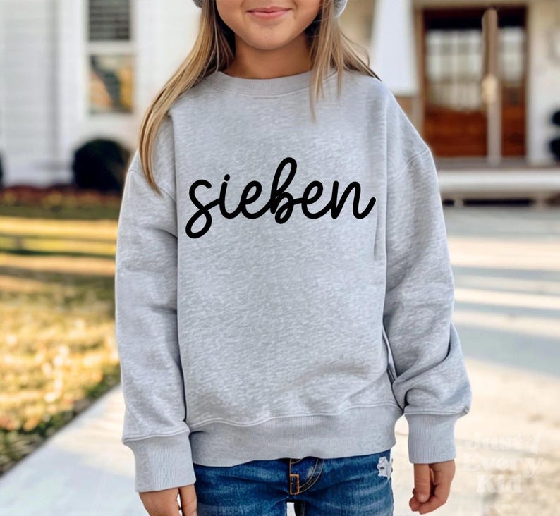May include: A light gray sweatshirt with the word "sieben" in black script. The sweatshirt has long sleeves and a crew neck. The person is wearing blue jeans.