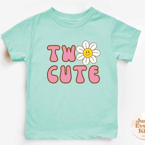 May include: A light blue toddler t-shirt with the words "TW" and "CUTE" in pink bubble letters with a brown outline. A white daisy with a yellow center replaces the "O" in "TWO". The shirt has short sleeves.