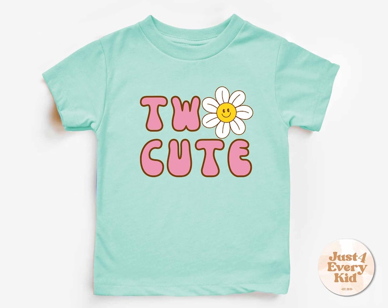 May include: A light blue toddler t-shirt with the words "TW" and "CUTE" in pink bubble letters with a brown outline. A white daisy with a yellow center replaces the "O" in "TWO". The shirt has short sleeves.
