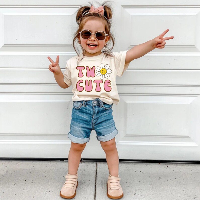 May include: A young child wearing a cream-colored t-shirt with the words "TWO CUTE" in pink and a daisy graphic. The child is also wearing blue denim shorts, sunglasses, and tan shoes. The child is smiling and making a peace sign with their fingers.