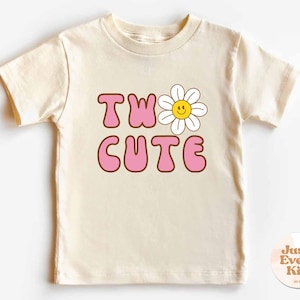 May include: Cream-colored short-sleeved t-shirt with the words "TWO CUTE" in pink bubble letters outlined in brown. A smiling daisy graphic is above the letter "O". The shirt is designed for children.