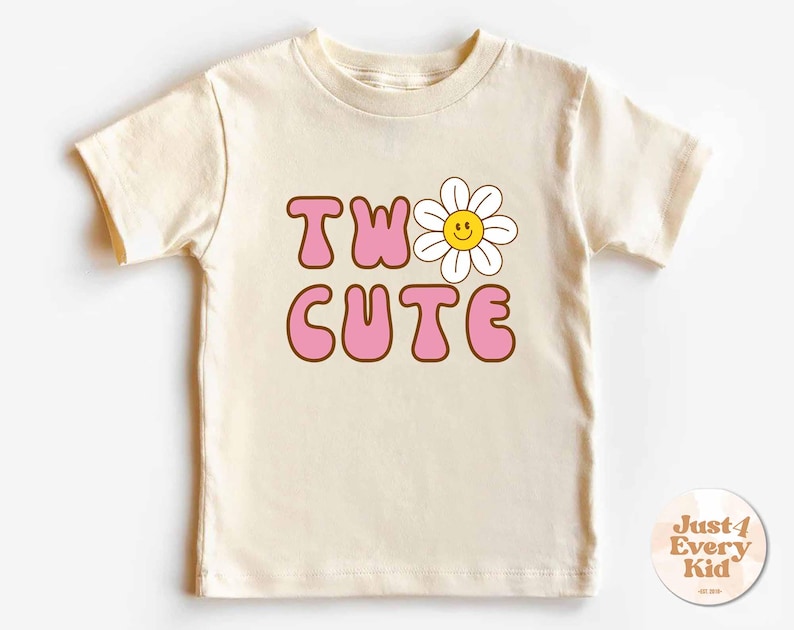 May include: Cream-colored short-sleeved t-shirt with the words "TWO CUTE" in pink bubble letters outlined in brown. A smiling daisy graphic is above the letter "O". The shirt is designed for children.