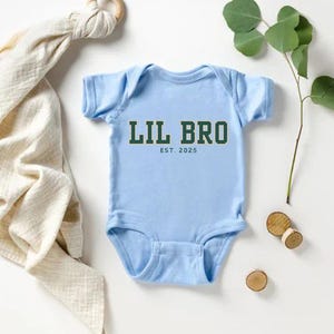 Puede incluir: Un body de beb&eacute; de manga corta en color azul claro con el texto LIL BRO en letras de bloque verde oscuro con un borde blanco, seguido por el texto EST. 2025 en una fuente verde oscuro m&aacute;s peque&ntilde;a centrada debajo del texto principal.