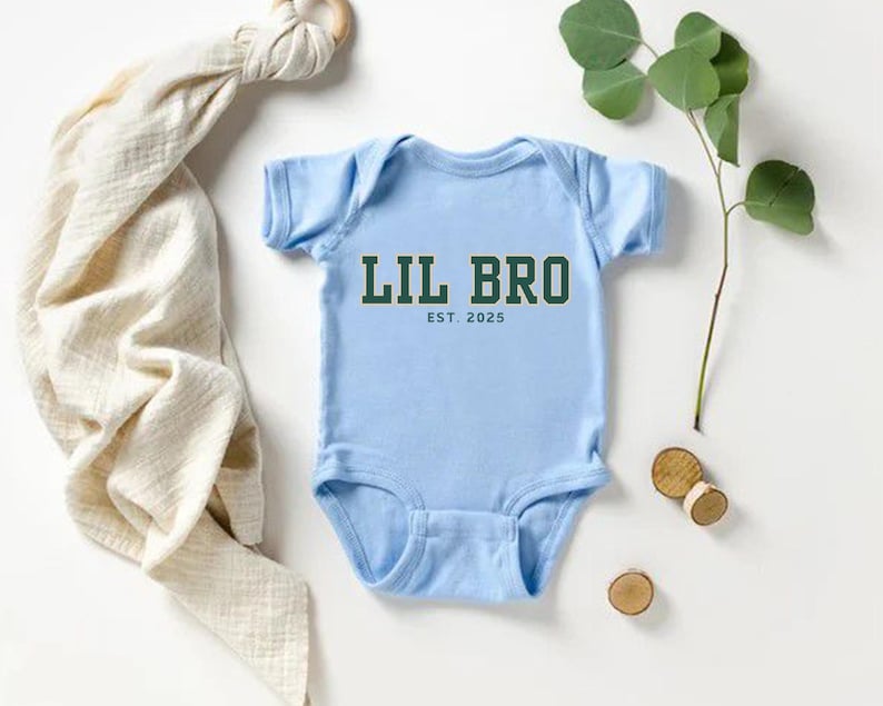Puede incluir: Un body de beb&eacute; de manga corta en color azul claro con el texto LIL BRO en letras de bloque verde oscuro con un borde blanco, seguido por el texto EST. 2025 en una fuente verde oscuro m&aacute;s peque&ntilde;a centrada debajo del texto principal.