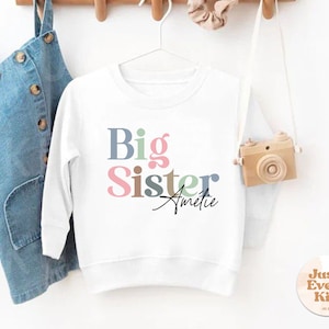 Puede incluir: Sudadera blanca de manga larga con el texto "Big Sister" en letras de colores y el nombre "Amelie" escrito en cursiva debajo. La sudadera está colgada en una percha con un vestido de mezclilla y una cámara de juguete de madera en el fondo.