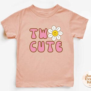May include: A light pink t-shirt with the words "TWO CUTE" in pink bubble letters with a brown outline. A white daisy with a yellow smiling face is above the word "CUTE".