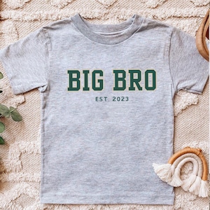 Puede incluir: Una camiseta de manga corta en color gris jaspeado con el texto en verde oscuro BIG BRO en una fuente de bloque en negrita, con el texto m&aacute;s peque&ntilde;o en verde oscuro EST. 2023 centrado directamente debajo de la inscripci&oacute;n principal.