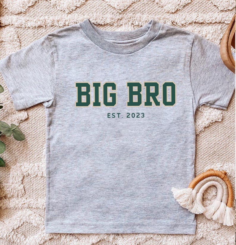 Puede incluir: Una camiseta de manga corta en color gris jaspeado con el texto en verde oscuro BIG BRO en una fuente de bloque en negrita, con el texto m&aacute;s peque&ntilde;o en verde oscuro EST. 2023 centrado directamente debajo de la inscripci&oacute;n principal.