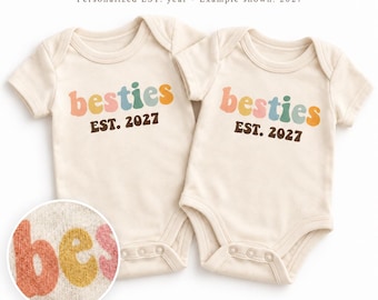 Op de afbeelding: Twee crèmekleurige babyrompertjes met korte mouwen. De tekst "besties" is gedrukt in veelkleurige, afgeronde letters boven de donkerbruine tekst "EST. 2027". Een cirkelvormige inzet toont een close-up van de getextureerde letters.