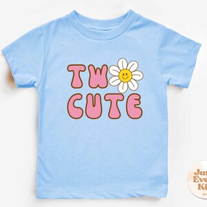 May include: A light blue toddler t-shirt with the words "TWO CUTE" in pink bubble letters with a brown outline. A white daisy with a yellow smiling face replaces the "O" in "TWO". The shirt has short sleeves and a crew neck.