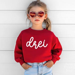 May include: A red sweatshirt with the word "drei" in white script. The child is wearing heart-shaped sunglasses and blue jeans. The sweatshirt has a crew neck and long sleeves.